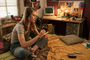Young woman sitting on bed, writing in notebook while listening to music. Room filled with personal...