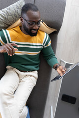 Smiling African-American man relaxing on a sofa, using a tablet, and enjoying leisure time at home in a comfortable and cozy environment.