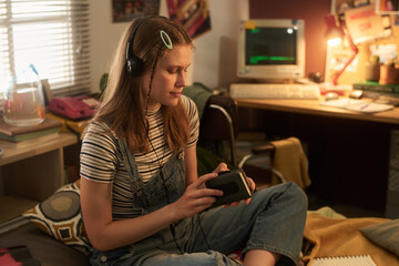 Young teenager listening to music with headphones in a retro-themed room with vintage decor and ambience, holding electronic device while sitting on chair © pressmaster