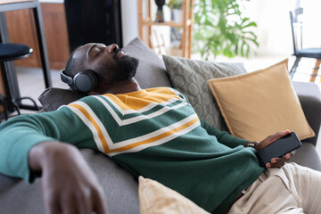 A joyful African-American man sits on a cozy couch wearing headphones, enjoying music or a podcast at home while holding his smartphone, relaxing in a casual and comfortable environment.