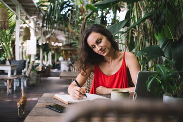 Woman in a red dress writing in a notebook while working on a laptop at an outdoor cafe surrounded by lush plants. A peaceful, tropical setting that inspires creativity and focus, cat walking nearby