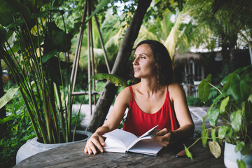 Woman in red dress reading a book outdoors, gazing thoughtfully into the distance. Surrounded by...