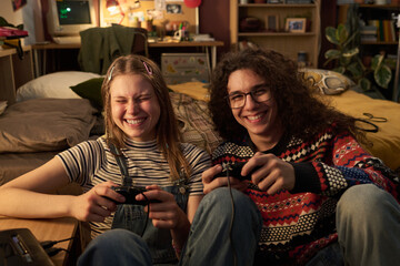 Smiling teenagers enjoying video games together while sitting in cozy living room setting filled...