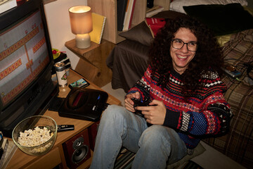 Smiling person with long curly hair sitting in front of TV while holding retro game controller,...
