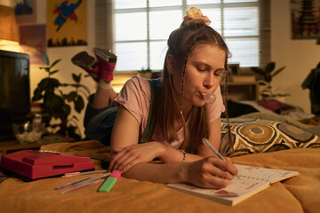 Young woman lying on bed and blowing bubble gum while writing in notebook during afternoon. Room...