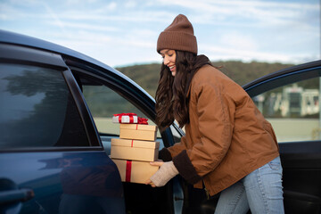 Woman putting wrapped Xmas gift boxes into car, delivering New Year presents. Lady enjoy atmosphere of holidays preparation and surprises