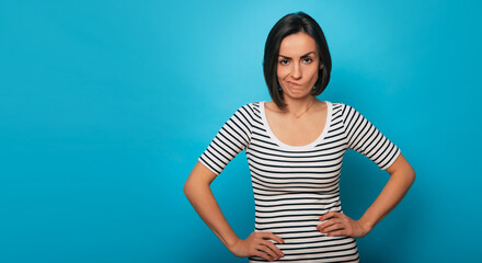 Young woman with short dark hair in striped shirt stands against a blue background, arms on hips, and wearing a humorous, unimpressed expression. Her body language suggests attitude and personality
