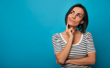 A young woman in a striped shirt stands against a blue background with a thoughtful expression,...