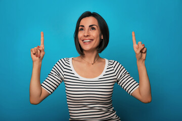Cheerful young woman in a striped shirt points both index fingers upward with an excited expression...