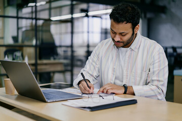 Obraz premium Focused ethnic man writing on paper while sitting at table with laptop and notepad