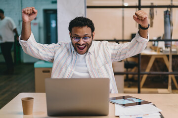 Excited ethnic man celebrating success with laptop in modern workspace