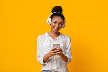 App For Music. Happy Afro Girl With Smartphone And Wireless Headphones Listening Her Favorite Songs And Lookin At Camera, Yellow Background