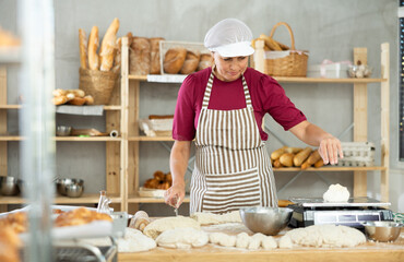 Adult female baker cutting dough with scraper and weighing pieces on scales