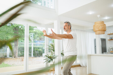 Mature man practicing focused Tai Chi poses in a modern, bright living space with large windows and greenery