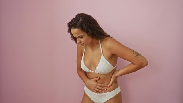 Painful stomach ache, young hispanic woman in bikini feeling ill and unwell over isolated pink background