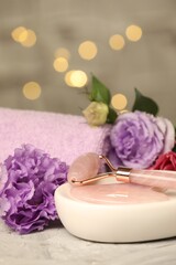 Face roller, gua sha tool, towel and beautiful flowers on gray textured table against blurred lights, closeup