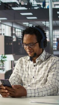 Vertical video African american general manager reviews forecasts while he listens to music on headset, feeling relaxed and multitasking at his desk. Data analyst focuses on objectives, strategy