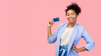 Shopaholic. Happy black woman holding credit card, resting hand on her hip. Copyspace, pink studio wall