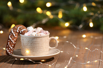 Cup of hot cocoa with gingerbread cookie and garland on wooden table against blurred Christmas background