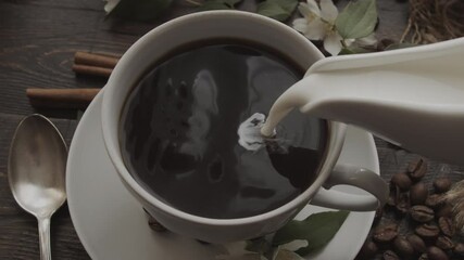 Pouring cream into black coffee in cup, with cinnamon sticks, coffee beans and flowers artfully arranged on rustic wooden table nearby. Close-up view - Powered by Adobe