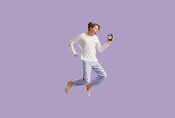 Shocked young man in pajamas with alarm clock jumping on lilac background