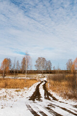 Autumn landscape. Sunny November day, the first white snow is melting on a field of dry grass. There are traces of cars on the brown dirt road. There is a cold blue sky above the horizon.
