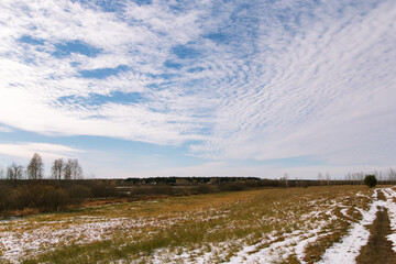 Blue cold sky with white clouds over a field with melting snow. Autumn landscape. Sunny November day.