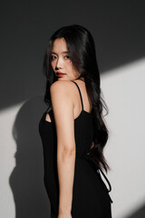 Fototapeta premium 光と影の背景で撮影をした黒いドレスを着た若い女性のシックなポートレート