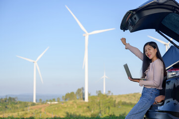 Asian woman working with her car trunk open and gazing at her laptop, content with a job well done in the midst of a wind farm that generates renewable energy
