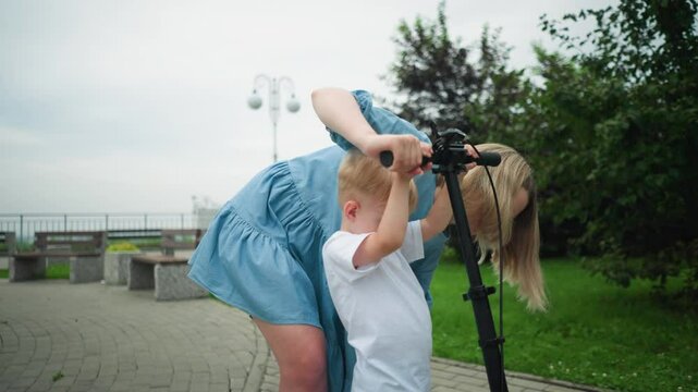 A mother carefully guides her son on how to stand properly on a scooter, they are standing together on an interlocked path as she helps him place his hands on the scooter's handlebar
