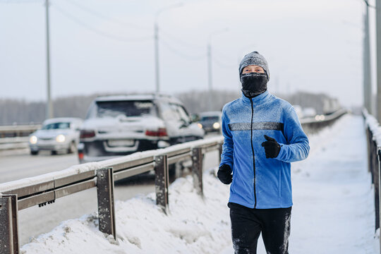 middle aged caucasian man runs on river bridge in winter, blue sport jacket, black trousers and balaclava to protect face from cold, active lifestyle concept, jogging in winter, sport life, copy space