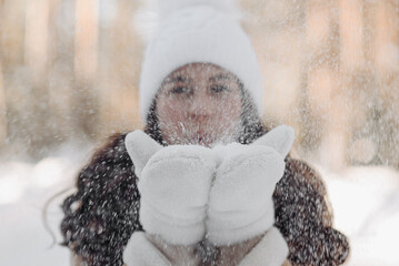 close-up portrait of young smiling caucasian girl in sunny day in winter snowy forest in white cap, beige fur jacket and mittens, blowing on snow in hands, snowflakes flying