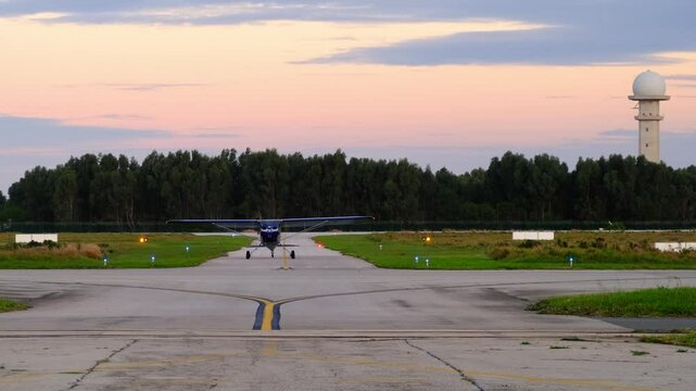 A small training plane taxiing towards camera on the taxiway after landing at the airport. Scenic sunset landscape with the forest and radar tower on the background