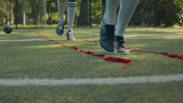 Low section of two male players wearing sports shoes and white socks performing speed and agility drill using flat ladder like training tool on grassy field during soccer training session with coach