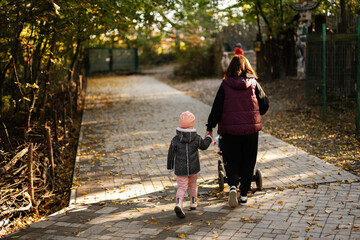 Fototapeta premium A mother and her child walk hand in hand with a stroller along a tree-lined path on a sunny autumn afternoon, creating a warm and serene family moment.