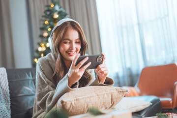 Happy asian woman wearing headphone enjoy playing game on smartphone at home near Christmas tree
