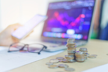 A stack of coins on a desk in front of an open laptop, with financial charts displayed on the...