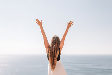 A woman stands on a rocky beach with her arms raised in the air. The sky is clear and the ocean is calm. The woman is happy and enjoying the beautiful scenery. © svetograph