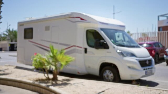 Blurry outdoor photo of a parked caravan in a sunny area featuring palm plants in the foreground and other vehicles in the background