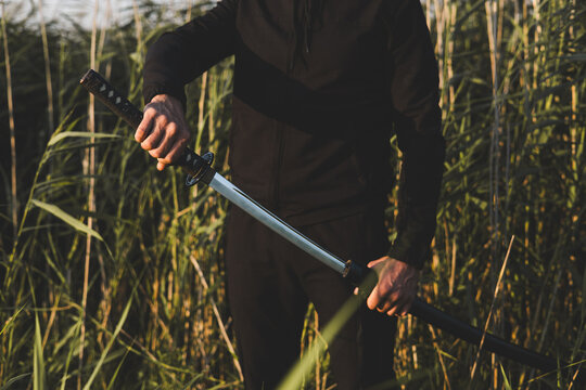 Close up of a martial artist unsheathing his katana sword in nature, surrounded by tall grass, with a focused and determined expression on his face. The man is training with a katana.