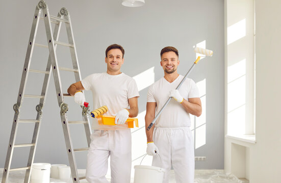 Portrait of smiling happy team of house painters looking at camera holding paint rollers and going to paint the wall of new apartment. Building contractors doing repair renovation preparing to move.