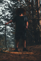 A masked swordsman stands on a path in the forest, holding a katana in his hand. The swordsman wears a straw hat and a black suit, stands proudly holding a katana and looks forward.