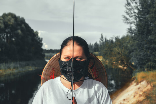 A woman stands by the river on a cloudy day, wearing a samurai mask and hat holding a katana with a serious expression mixing Japanese culture and tradition in a captivating scene. Woman with katana.