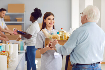 Portrait of a young cheerful woman volunteer working at charity center and giving cardboard donation food box for mature man in charitable foundation. Humanitarian aid and volunteering concept.