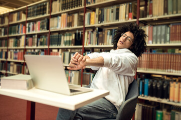 Young man sitting at the laptop in the library and looking tired