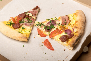Close-up of a pizza slice with ham, sausage, tomatoes, and herbs, placed on a parchment-covered surface. A bite is taken from the crust beside it.