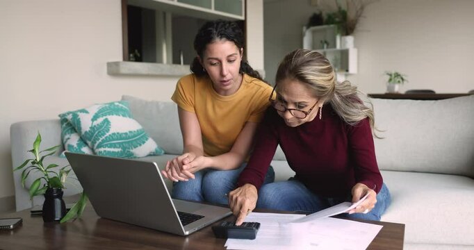 Mature senior mother teaching young adult daughter to pay domestic bills, check paper invoices on calculator, using online financial application on laptop, giving high five to grown child
