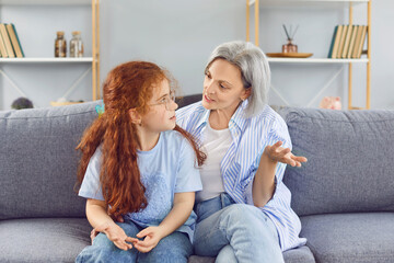 Obraz premium Portrait of a gray-haired senior grandmother sitting on sofa at home with her little granddaughter child girl and talking in the living room. Family leisure and generations concept.