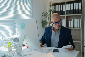 Focused mature manager using calculator and working on desktop computer, checking financial documents and calculating taxes in modern office