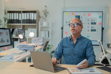 Fototapeta premium Businessman is sitting at his desk in the office, working on a laptop and reviewing financial documents, analyzing charts and graphs, and developing a new marketing strategy